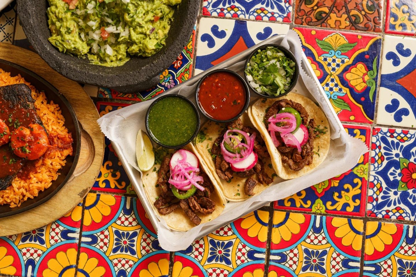 Tacos garnis d’oignons roses, sauces, guacamole et riz sur une table aux carreaux colorés