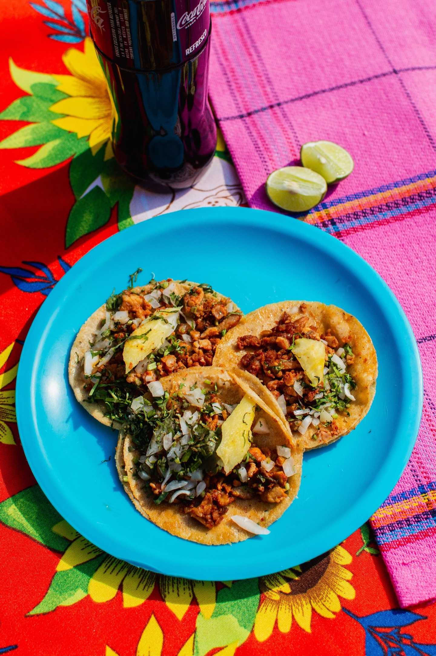 Assiette bleue de tacos garnis de viande, d’ananas et d’oignons, sur une nappe colorée